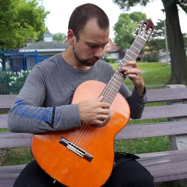 Brett holding a classical guitar wearing a grey shirt and playing in a park.