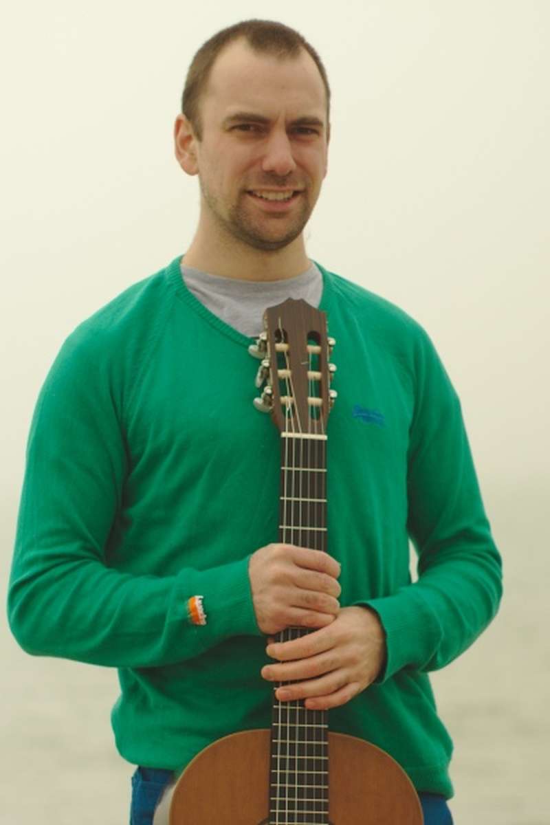 Brett Vachon holding a guitar at the beach wearing a green sweater